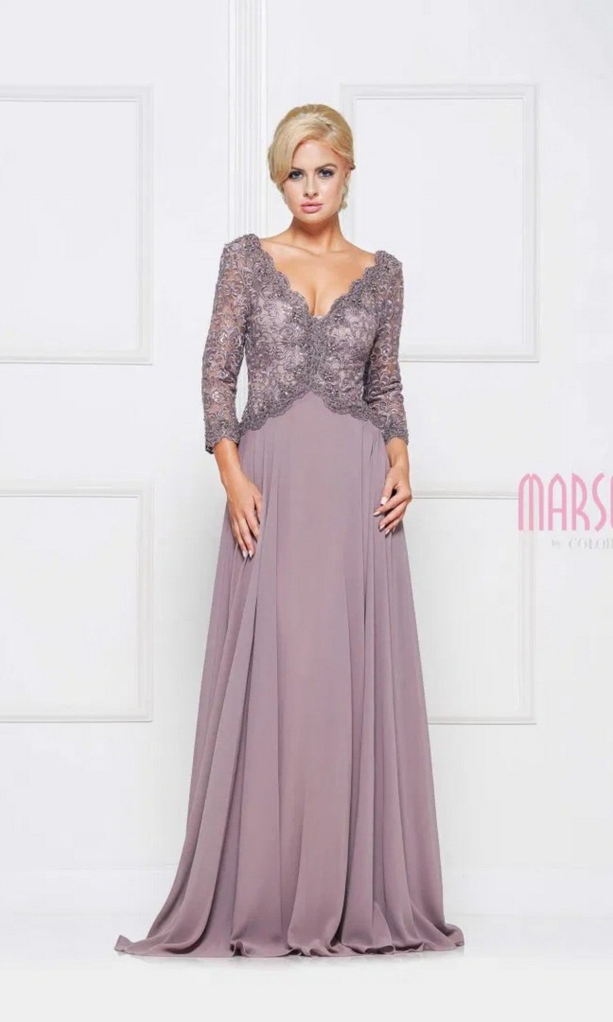 A woman stands against a white paneled background wearing the Marsoni M225 long taupe formal dress, featuring a lace-embroidered bodice, three-quarter sleeves, and a flowing chiffon A-line skirt.